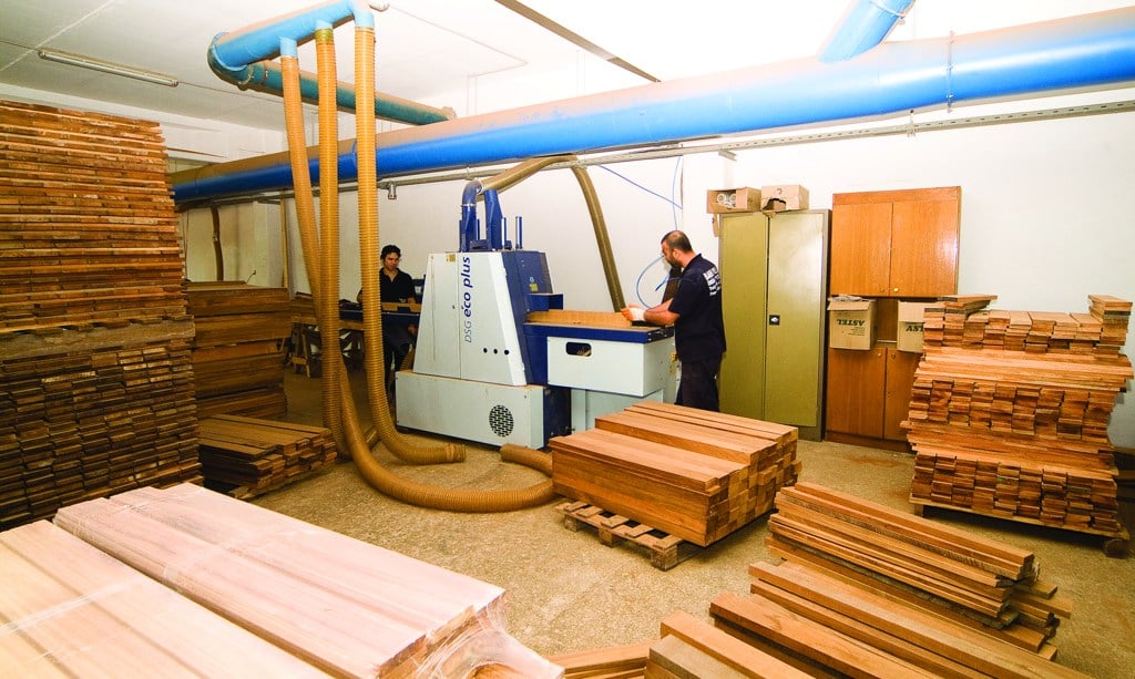 Operators at production machinery surrounded by timber stacks and dust extraction pipes